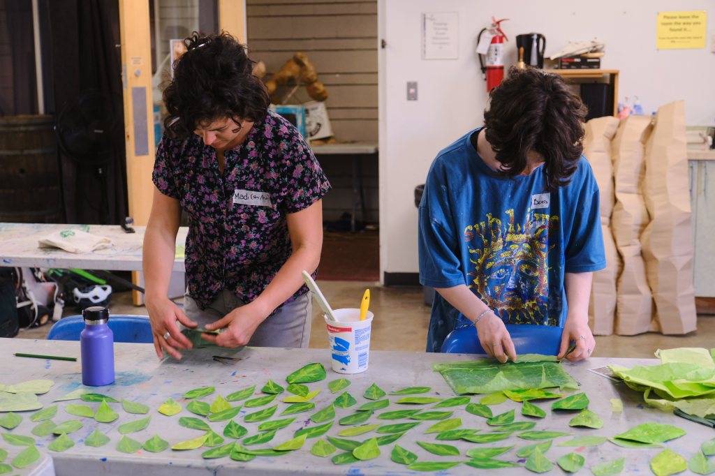 Two people lean over a table to work on a collection of green paper leaves.