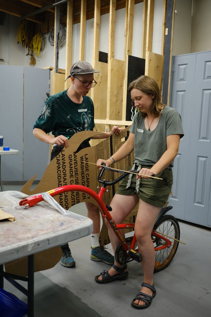 One person sits on top of a loose bike trailer, attached on one end to a craft table, while another holds a cardboard cutout of flames alongside.