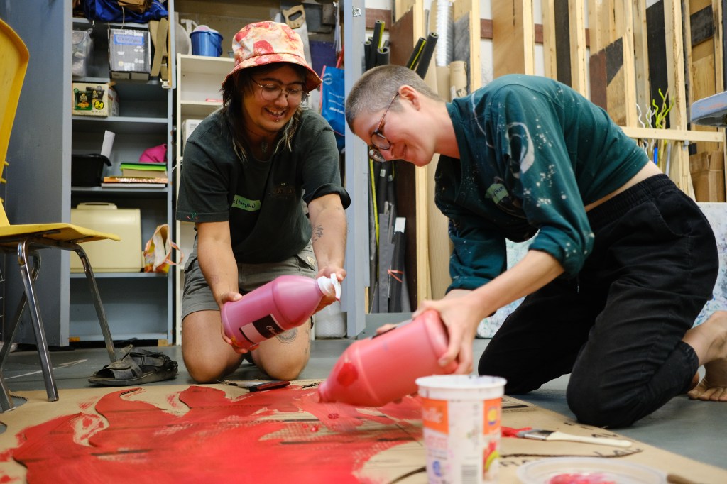 Two people smile while kneeling on the ground painting a piece of fire-cardboard red.