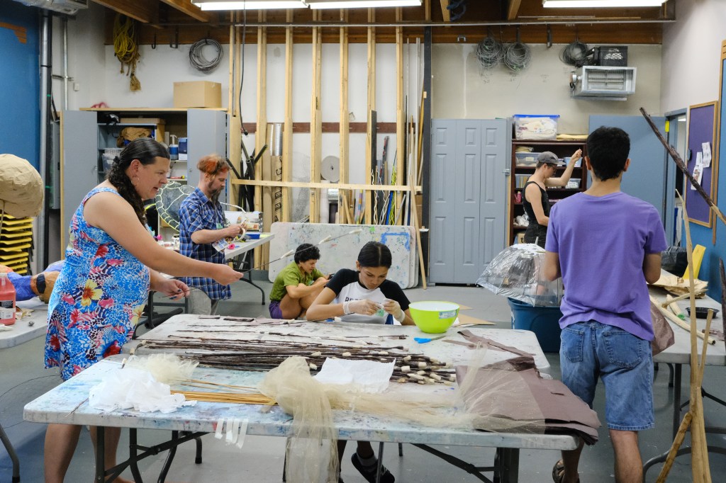 Six people in a room full of giant puppet crafts, all engaged in their tasks. One sits on the floor, and one in a chair, the rest stand.